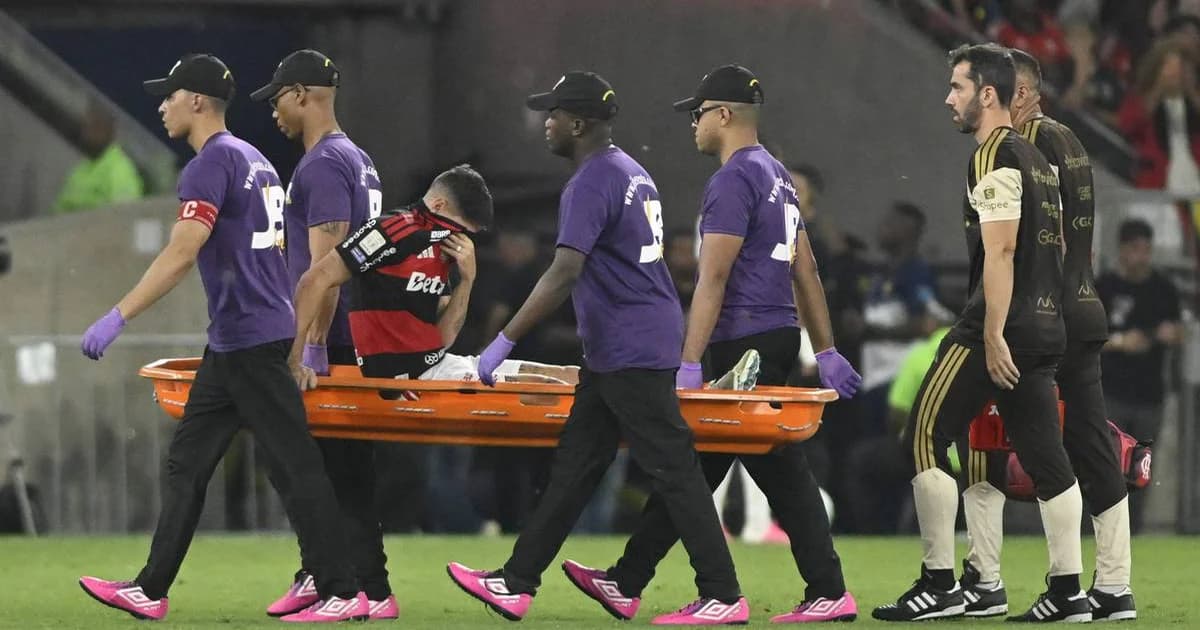 Lateral-esquerdo com lesão muscular segurando a parte posterior da coxa no gramado; equipe médica e estádio ao fundo