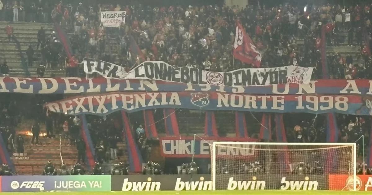 Estádio com gramado coberto por fumaça, sinalizadores e objetos; partida cancelada após invasão, torcida em silhuetas.