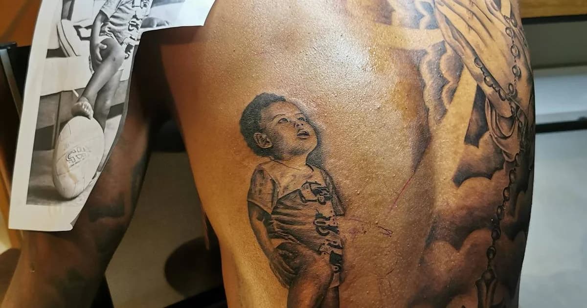 Tatuador do Flamengo tatuando o braço de um jogador, estúdio com janela mostrando estádio iluminado e bandeiras abstratas em vermelho e preto.