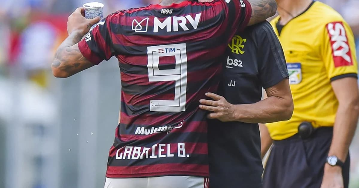 Treinador e atacante se reencontram no campo com torcida em vermelho e preto celebrando, atmosfera de vitória no estádio.