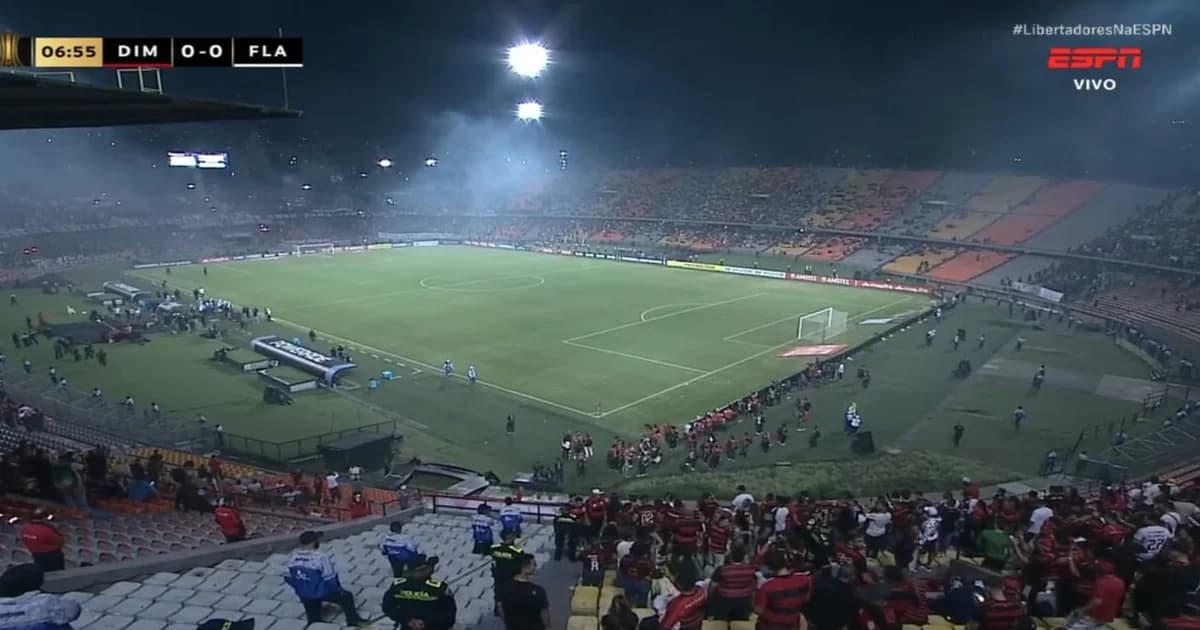 Cena no Estádio Atanasio Girardot: jogo do Flamengo pela Libertadores interrompido, árbitro sinaliza, torcida em tumulto e fumaça.