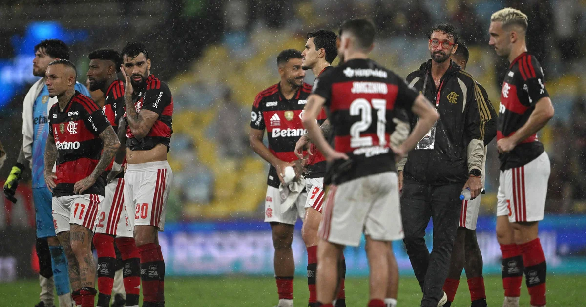 Estádio com arquibancadas vermelhas e pretas, técnico em silhueta abatido e duas taças caídas, simbolizando crise e derrotas do Flamengo.