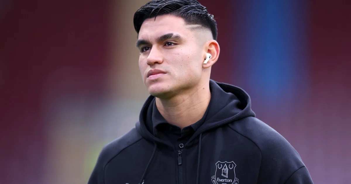 Jogador reserva no banco de um estádio inglês, torcida vibrante em azul, vermelho-preto e preto-branco ao fundo