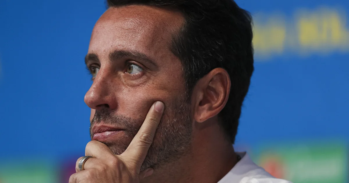 Executivo de costas com celular em estádio, bandeiras vermelho e preto, imprensa reunida e clima de confirmação da procura.