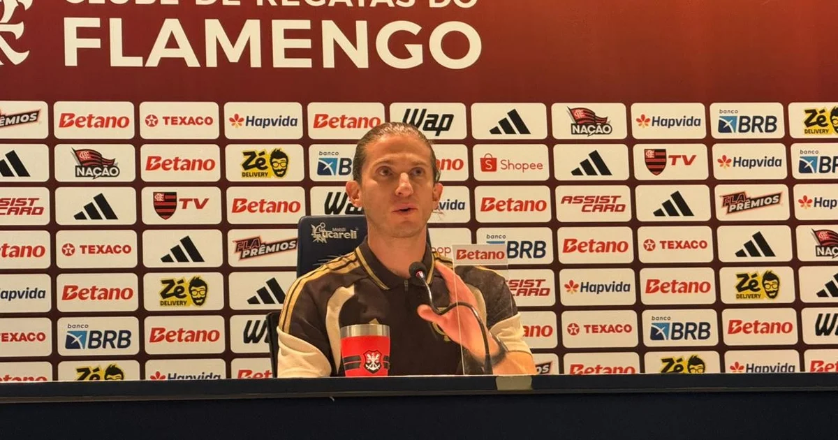 Treinador em coletiva no estádio após vitória 3 a 0, torcida com faixas "Não ao racismo" e clima de apoio, cores vermelho e preto.