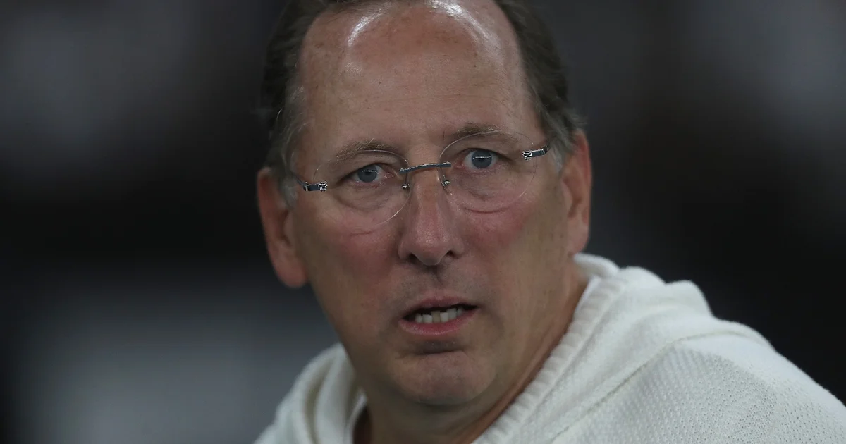 Treinador de costas observa estádio dividido por faixas em preto-e-branco (Botafogo) e vermelho-e-preto (Flamengo), torcida ao fundo, clima tenso
