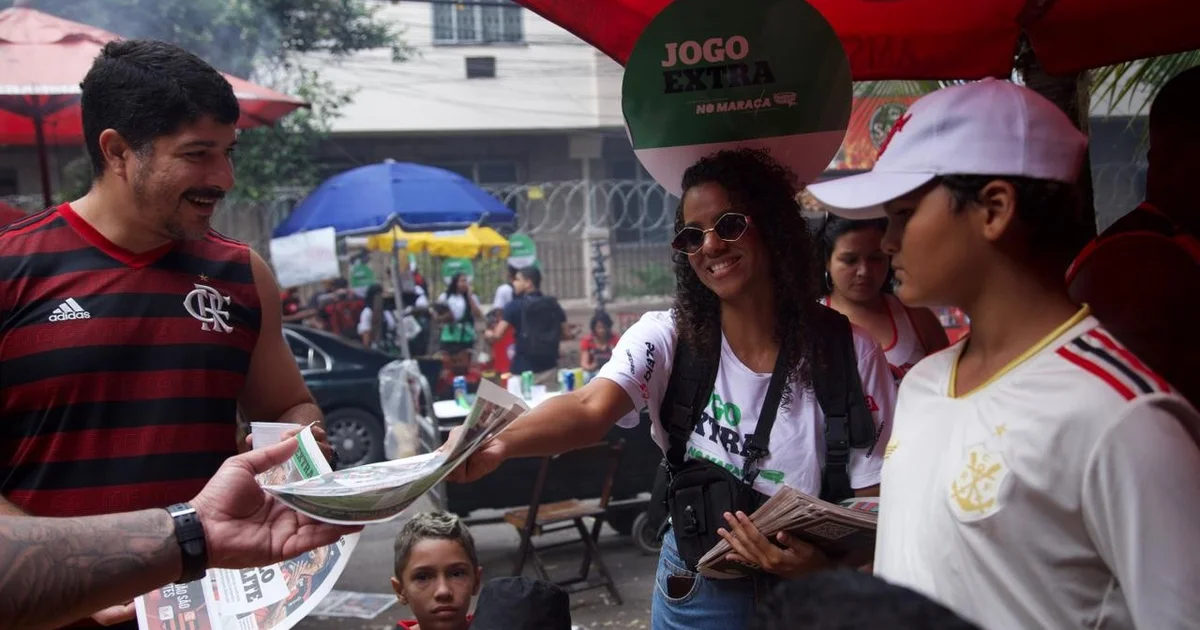 Torcedores no entorno do Maracanã recebendo cadernos e copos promocionais antes do Fla‑Flu, multidão e atmosfera de estádio.