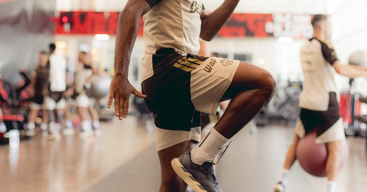 Ilustração editorial: treino no estádio ao entardecer, equipe médica cuidando de jogador e comissão técnica com jogo remarcado.