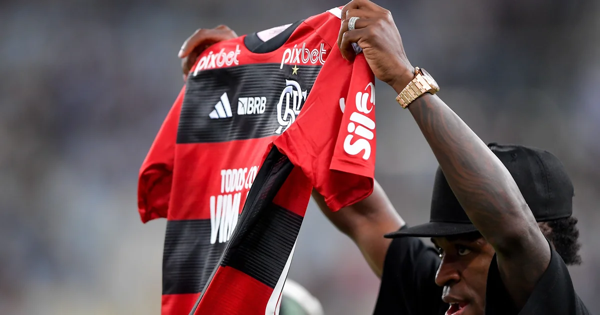 Estádio noturno com torcida em vermelho (Flamengo) e vinho (Lanús), placar 0-1, jogadores em silhueta; tensão e menção à denúncia de racismo.