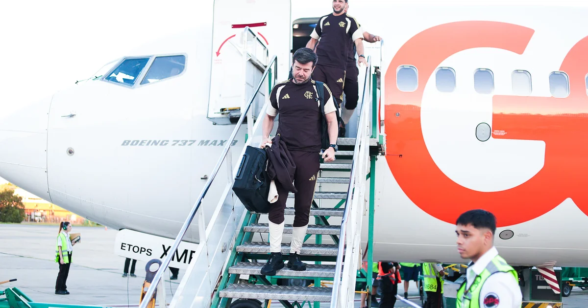 Aeronave exclusiva do Flamengo ao anoitecer ao lado do estádio, equipe carregando equipamentos; bola e arquibancada iluminada ao fundo