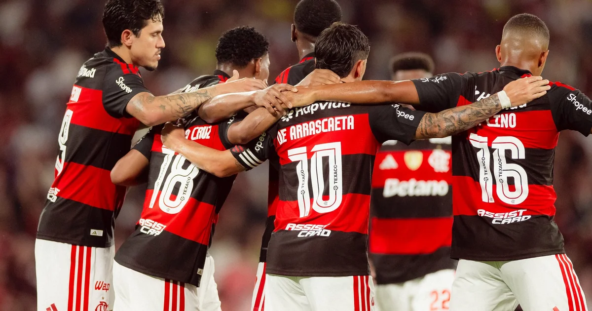 Estádio em comemoração (vermelho/preto) à esquerda; CT à direita, clima frio, jogadores afastados e tensão no Flamengo.