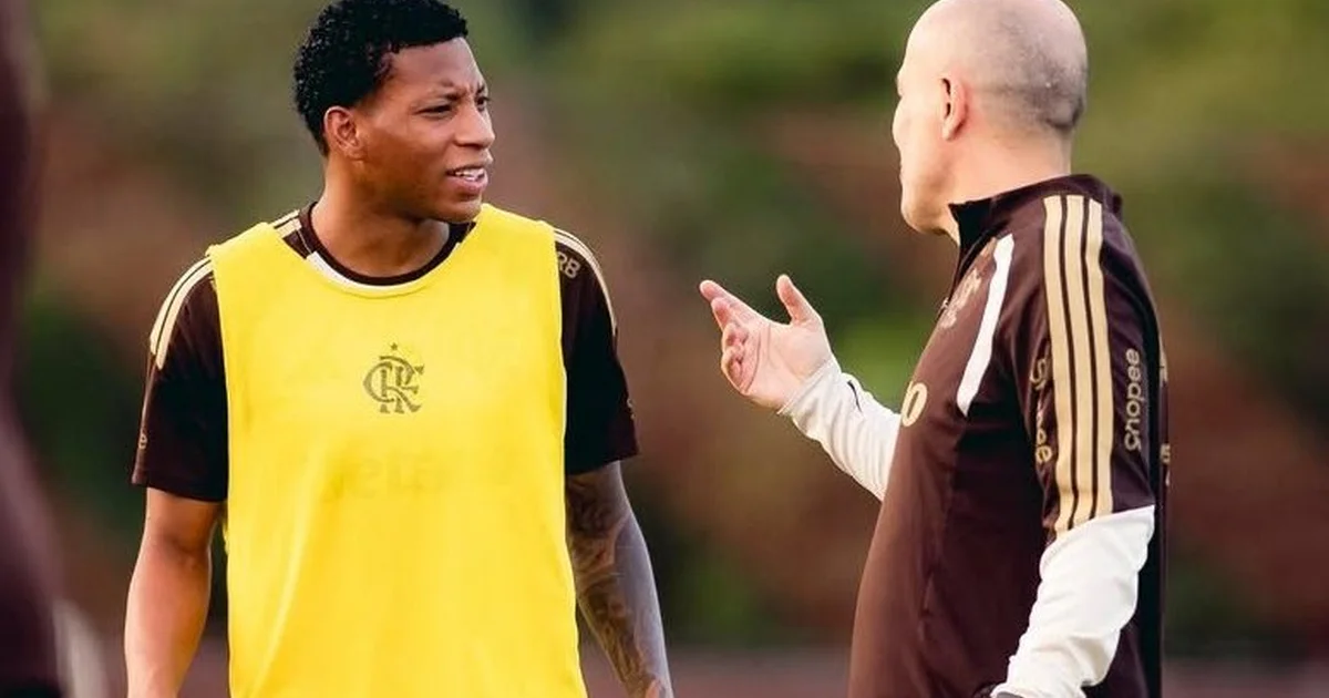 Jogador anônimo consolado por companheiros da seleção em estádio dividido entre pressão sombria do clube e luz solar, simbolizando alívio e união.