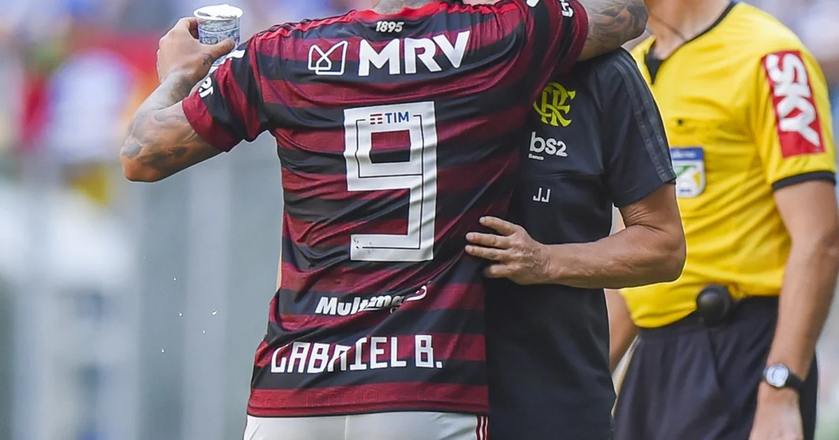 Treinador e atacante se reencontram no campo com torcida em vermelho e preto celebrando, atmosfera de vitória no estádio.