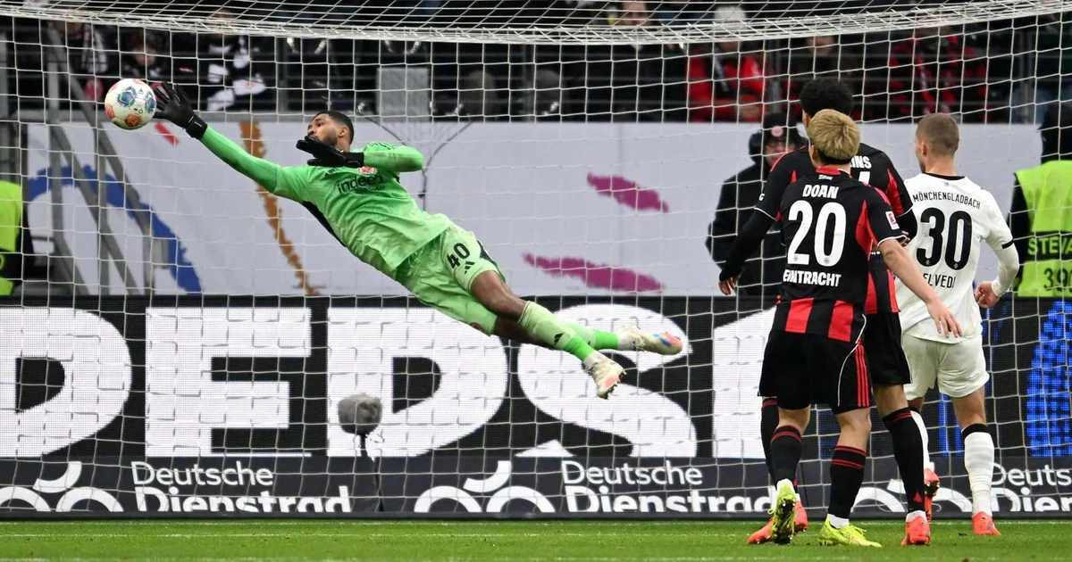 Goleiro jovem lança passe longo em estádio iluminado; defesa decisiva, torcida ao fundo e clima de vitória.
