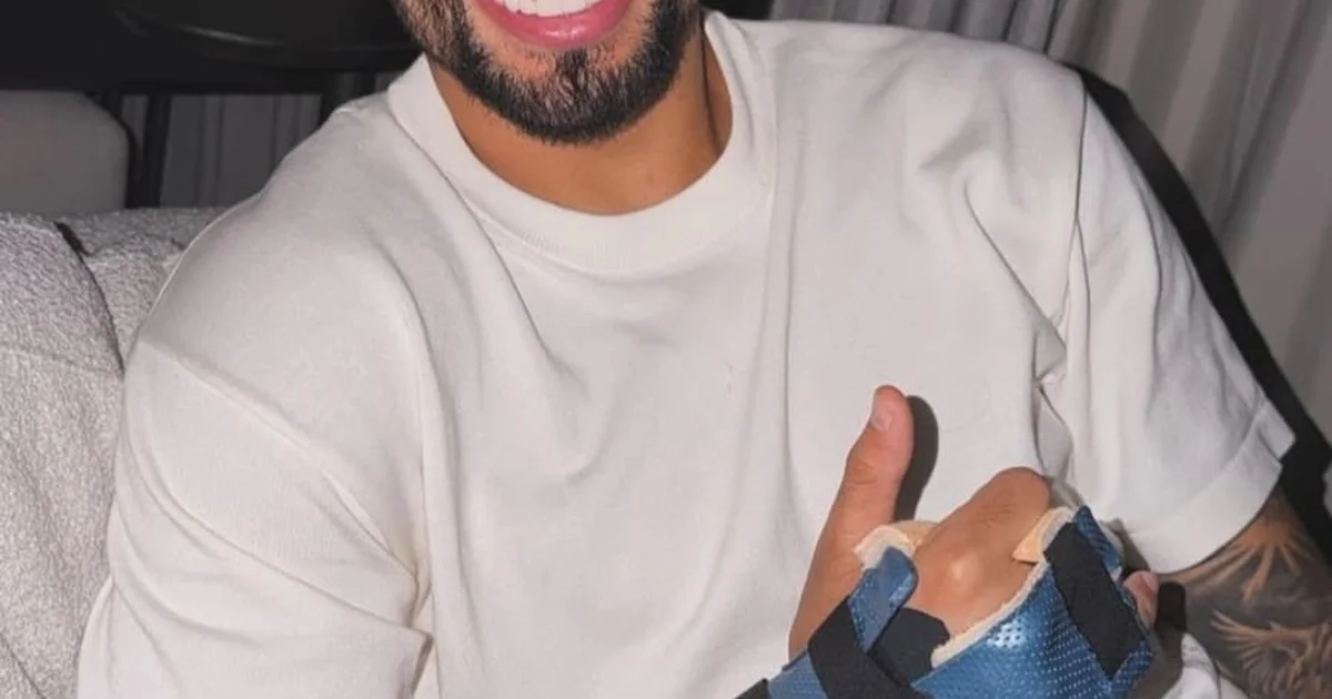 Jogador com mão imobilizada usando proteção treina e participa de clássico em estádio lotado, com torcida e iluminação intensa.