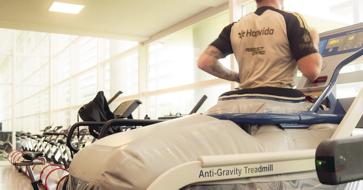 Silhueta em esteira antigravidade no estádio, equipe médica ao lado, recuperação do tornozelo, bandeiras vermelhas e pretas